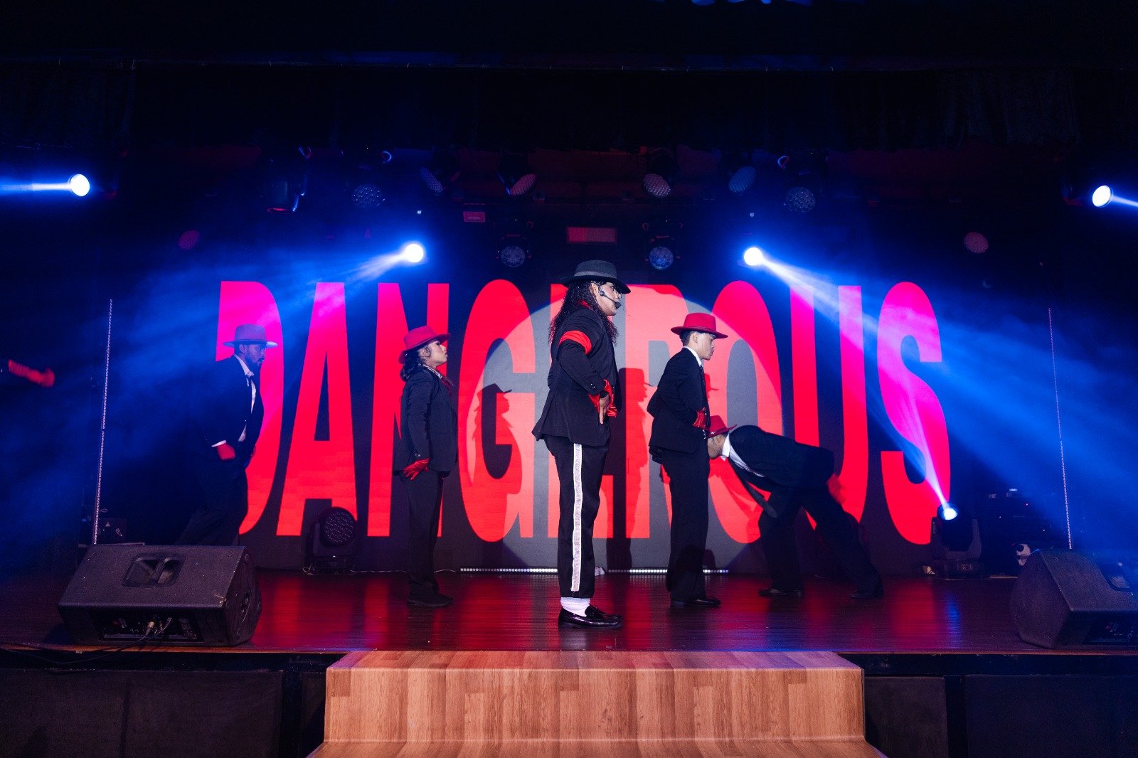 Full company on stage in front of a giant red DANGEROUS backdrop under blue spotlights