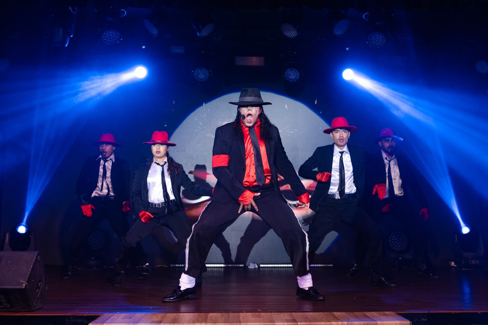 Michael Jackson tribute lead in a black fedora under a spotlight, dancers in red hats behind him